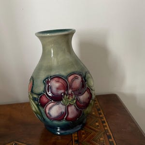 Könnte beinhalten: Eine Keramikvase mit bauchigem Körper und ausgestelltem Hals, verziert mit einem handgemalten Motiv einer großen, dunkelroten Blume mit weißen Akzenten auf einem gedämpften salbeigrünen Hintergrund, ergänzt durch grüne Blätter und einen dunkelblauen Fuß.