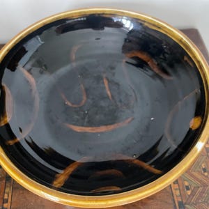 May include: A round ceramic bowl with a glossy black interior featuring abstract, curved orange-brown brushstroke patterns. The bowl has a thick, smooth rim finished in a warm, golden-brown glaze that contrasts with the dark centre basin.