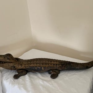 May include: A wooden alligator sculpture with a detailed, textured body and open mouth. The alligator is painted in shades of brown and black, with visible wear and tear. The sculpture is resting on a white surface.