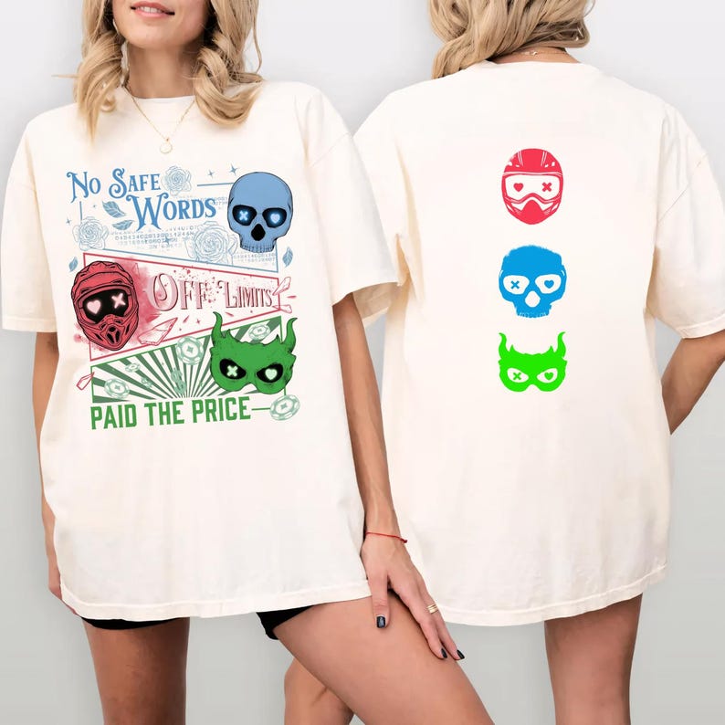 May include: Cream-colored t-shirt featuring graphic designs on the front and back. The front displays a blue skull, a red helmet, and a green mask with text that reads "No Safe Words", "Off Limits", and "Paid the Price". The back has three colorful skull designs.