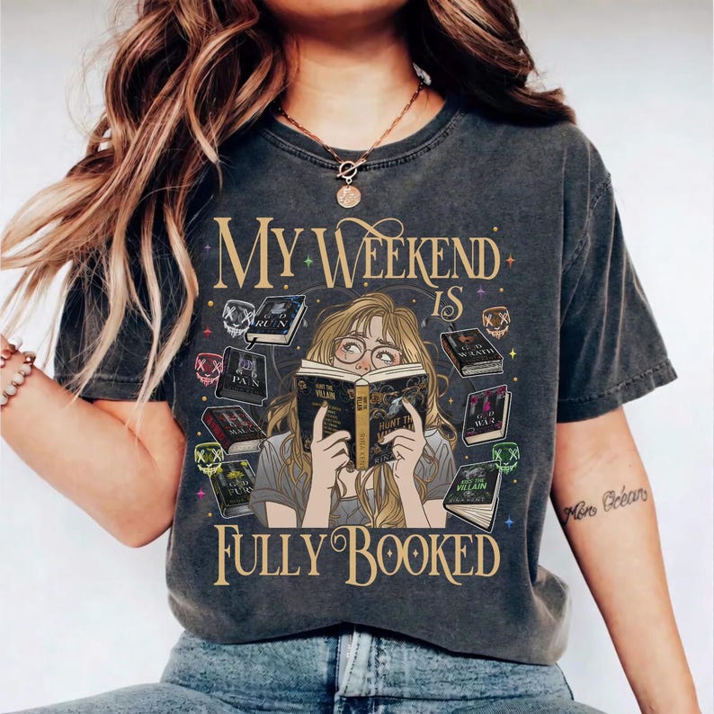 Pu&ograve; includere: T-shirt grigio scuro con la scritta "My Weekend Is Fully Booked". La grafica presenta un personaggio dei cartoni animati che legge un libro, circondato da altri libri. La maglietta ha un aspetto vintage e usurato.