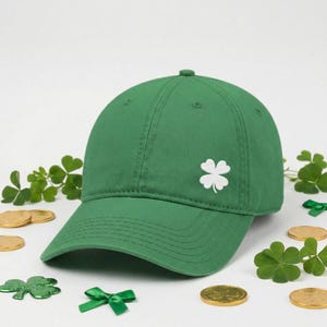 以下が含まれることがあります： 白い四つ葉のクローバーのエンブレムが付いた緑色の野球帽。画像には、シャムロック、金貨、緑のリボンが含まれており、聖パトリックの日のテーマを連想させます。