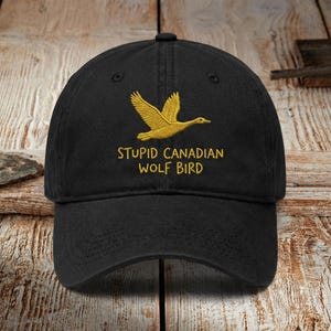 Könnte beinhalten: Schwarze Baseballkappe mit einem goldgestickten fliegenden Vogel und dem Text "STUPID CANADIAN WOLF BIRD". Die Kappe hat einen gebogenen Schirm und einen verstellbaren Riemen. Der Hut ist aus einem weichen, strapazierfähigen Stoff gefertigt.