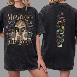 Pu&ograve; includere: T-shirt nero con la scritta "My Weekend Is Fully Booked" e una grafica di una persona che legge circondata da libri. Il retro della maglietta presenta una pila verticale di libri illustrati.