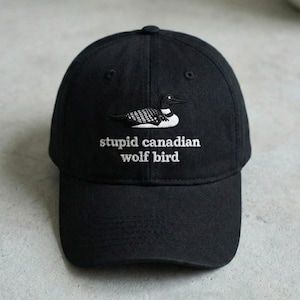 Peut inclure: Casquette de baseball noire avec un oiseau brodé blanc et le texte "stupid canadian wolf bird". La casquette a une visière incurvée et un bouton sur le dessus. Le chapeau est en tissu doux.