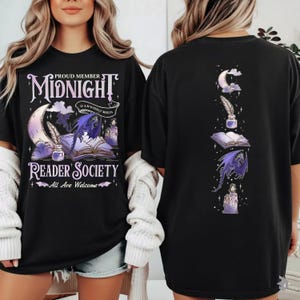 Op de afbeelding: Zwart t-shirt met een afbeelding van een maan, veer, open boek en paarse draak. De tekst luidt "Proud Member Midnight Reader Society. All Are Welcome." De achterkant heeft een vergelijkbaar ontwerp.