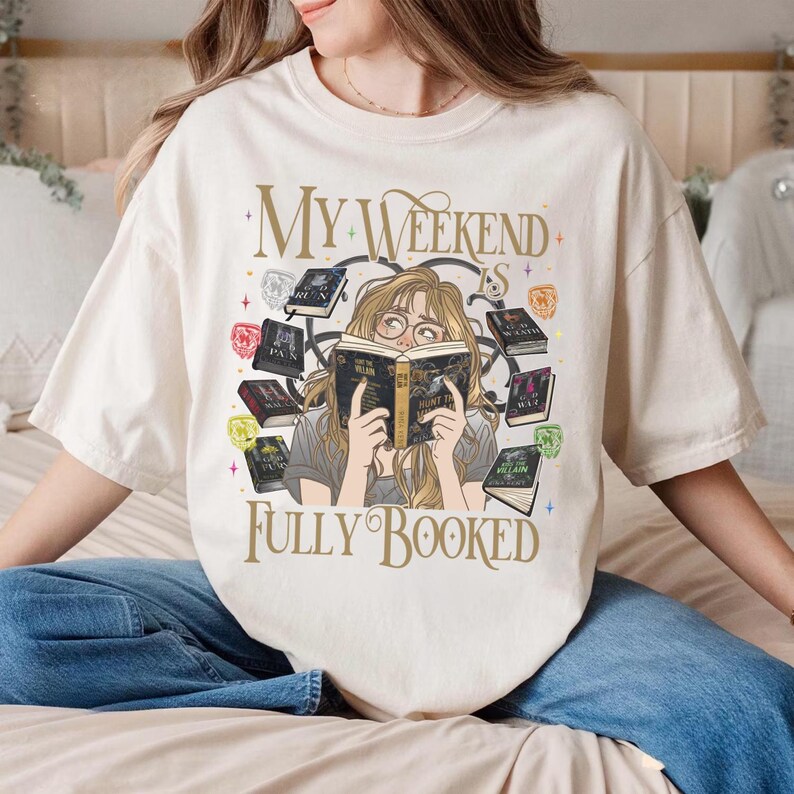 Pu&ograve; includere: T-shirt color crema con la scritta dorata "MY WEEKEND IS FULLY BOOKED". La grafica raffigura una donna che legge un libro, circondata da libri fluttuanti ed elementi decorativi.