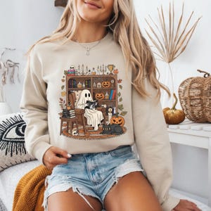 May include: A beige crewneck sweatshirt featuring a graphic print of a ghost sitting in an armchair reading a book. The illustration includes a bookshelf filled with books, pumpkins, candles, a black cat, and autumn leaves in shades of orange and brown.
