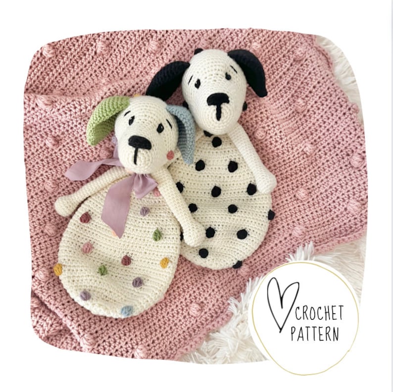 May include: Two crocheted dog plushies on a pink crocheted blanket. One dog is white with black polka dots and the other is white with multi-colored polka dots. The blanket has a textured, bumpy surface.