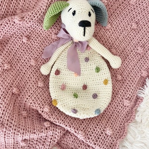 May include: A white crocheted dog toy with colorful polka dots. The toy has a purple ribbon around its neck and is lying on a pink crocheted blanket.