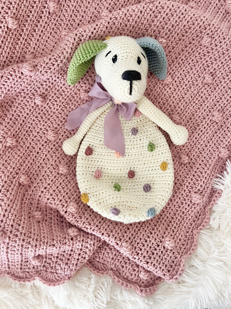 May include: A white crocheted dog toy with colorful polka dots. The toy has a purple ribbon around its neck and is lying on a pink crocheted blanket.