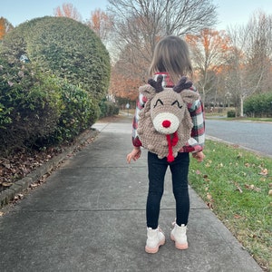 May include: A child wearing a handmade, tan-colored reindeer backpack with a red nose and brown antlers. The child is wearing a red and black plaid shirt, black pants, and white boots with pink soles, walking on a sidewalk.