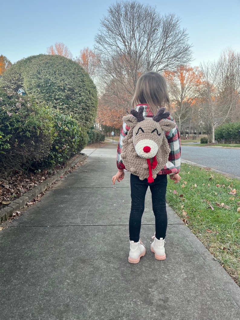 May include: A child wearing a handmade, tan-colored reindeer backpack with a red nose and brown antlers. The child is wearing a red and black plaid shirt, black pants, and white boots with pink soles, walking on a sidewalk.