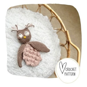 May include: A crocheted owl toy in brown and pink, with a yellow beak and white eyes, resting on a white textured surface. The image includes a circular logo with the words "Crochet Pattern".