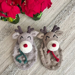 May include: Two handmade crochet reindeer Christmas ornaments. One is light gray with a green scarf, and the other is tan with a red scarf. Both have white noses with a red center, and dark brown antlers. Red poinsettias are in the background.