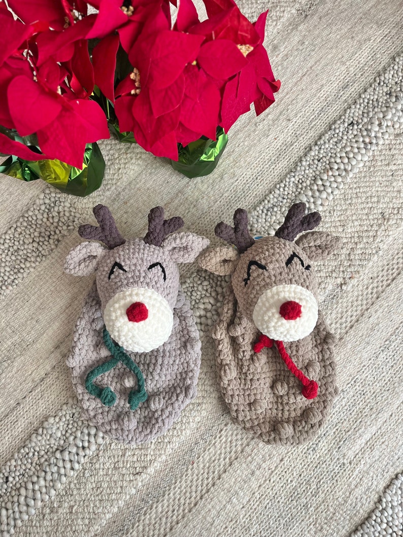 May include: Two handmade crochet reindeer Christmas ornaments. One is light gray with a green scarf, and the other is tan with a red scarf. Both have white noses with a red center, and dark brown antlers. Red poinsettias are in the background.