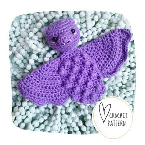 May include: A handmade purple crochet bat with textured wings and a smiling face. The bat is lying on a light blue and white textured surface. A white circle with a heart and the words "CROCHET PATTERN" is in the lower right corner.