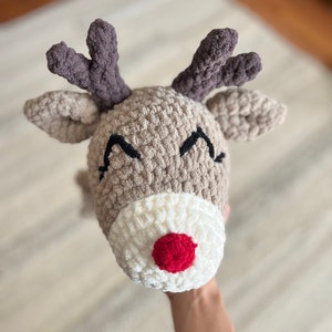 May include: A handmade, crocheted reindeer hat with a tan head, white muzzle, and a bright red nose. The hat features dark gray antlers and black stitched eyes, perfect for holiday festivities.