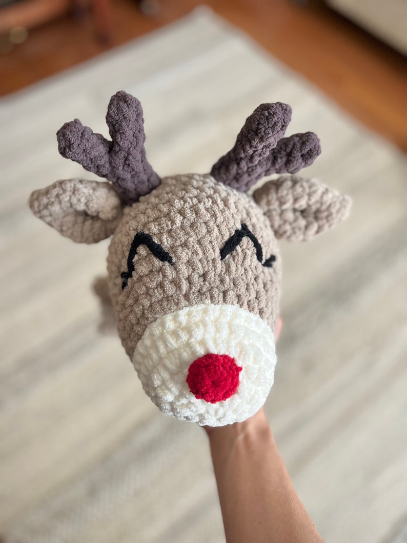 May include: A handmade, crocheted reindeer hat with a tan head, white muzzle, and a bright red nose. The hat features dark gray antlers and black stitched eyes, perfect for holiday festivities.