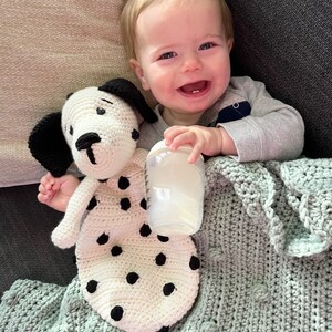 May include: A baby is lying on a gray blanket with a crocheted white and black spotted dog toy and a clear bottle. The baby is smiling and holding the bottle.