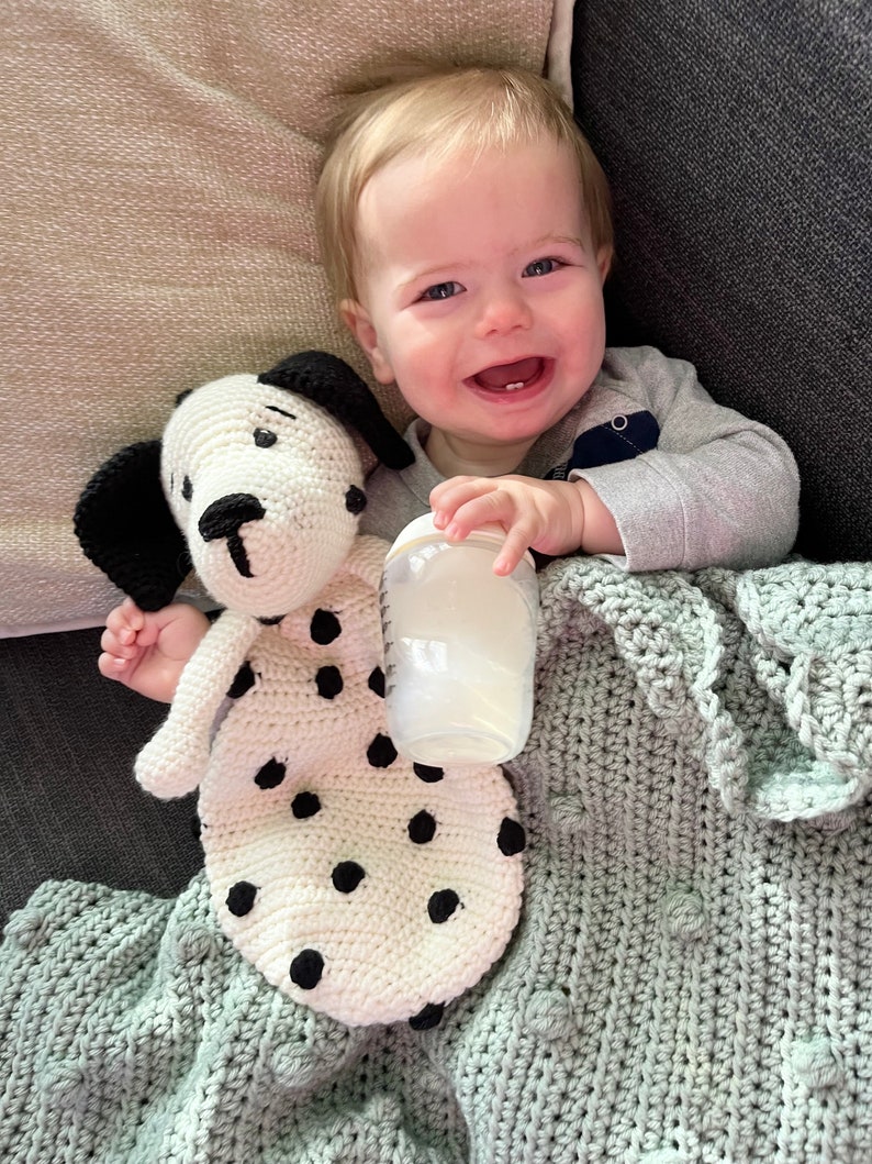 May include: A baby is lying on a gray blanket with a crocheted white and black spotted dog toy and a clear bottle. The baby is smiling and holding the bottle.