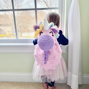 May include: A young child wearing a pink tutu and a crocheted unicorn backpack. The backpack is pink, purple, and yellow with a white horn.