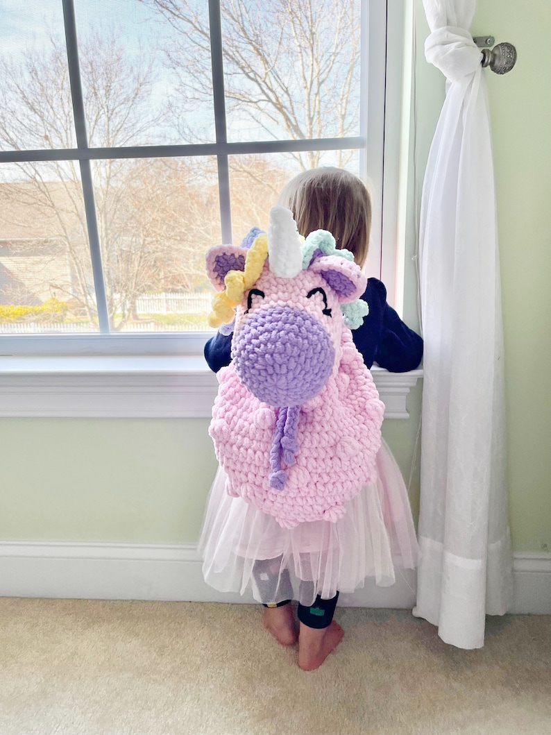 May include: A young child wearing a pink tutu and a crocheted unicorn backpack. The backpack is pink, purple, and yellow with a white horn.