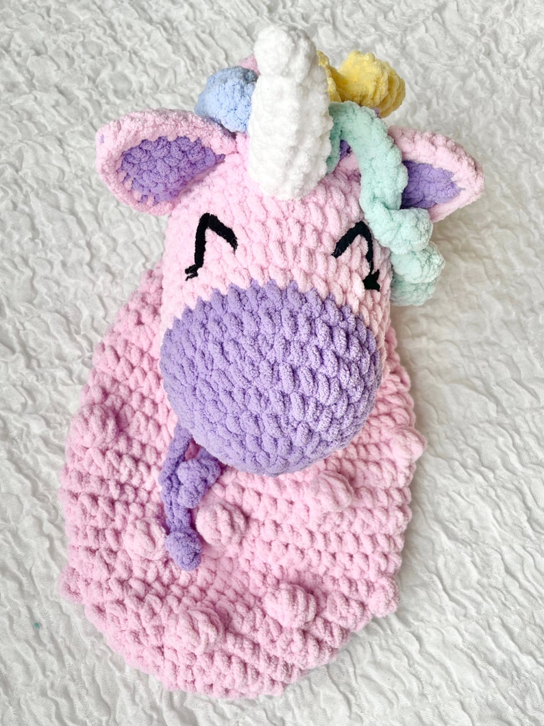 May include: A crocheted unicorn head with a pink and purple body, white horn, and rainbow colored mane.