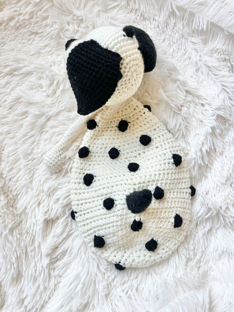 May include: A white crocheted stuffed animal shaped like a Dalmatian dog with black spots. The dog has a black crocheted nose and ears.