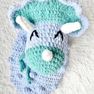 May include: A crocheted plush toy shaped like a triceratops dinosaur. The dinosaur is light blue and green with white horns and eyes.