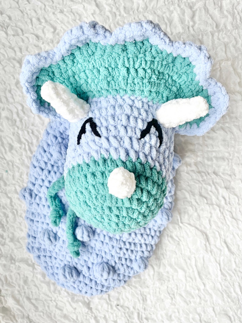 May include: A crocheted plush toy shaped like a triceratops dinosaur. The dinosaur is light blue and green with white horns and eyes.