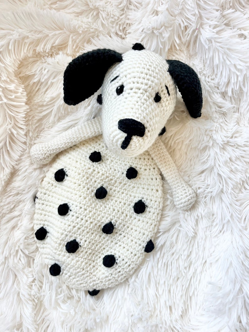 May include: White crocheted dog toy with black spots and ears. The toy is lying on a white fluffy surface.