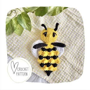 May include: A handmade crochet bee with yellow and black stripes, white wings, and black antennae. The bee is lying on a yellow and white checkered surface. The image includes the text "Crochet Pattern" inside a heart.