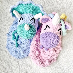 May include: Two crocheted stuffed toys, a green and blue triceratops and a pink and purple unicorn, with embroidered eyes and smiling expressions.