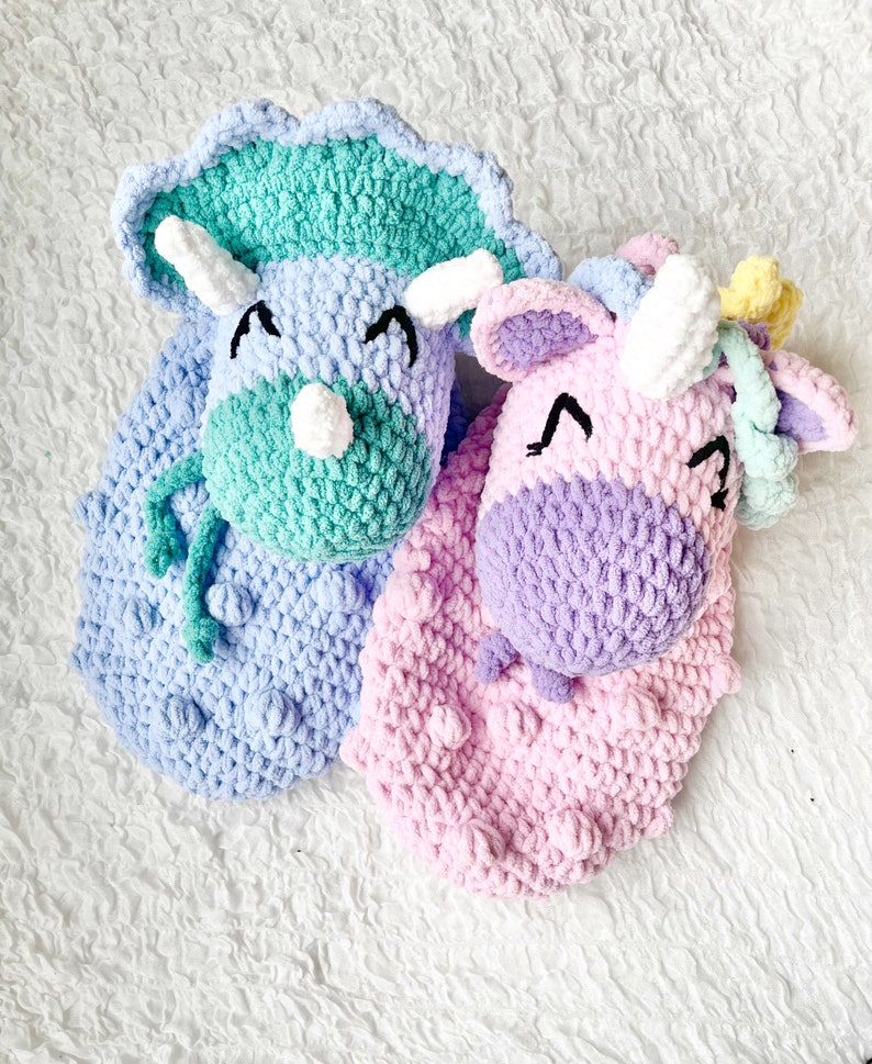 May include: Two crocheted stuffed toys, a green and blue triceratops and a pink and purple unicorn, with embroidered eyes and smiling expressions.