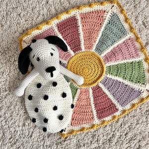 May include: A crocheted sunburst blanket in shades of pink, green, and yellow with a white crocheted dog with black spots lying on top.