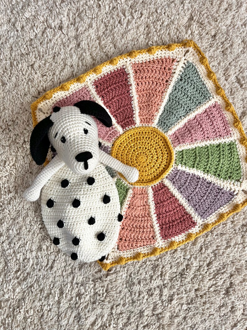 May include: A crocheted sunburst blanket in shades of pink, green, and yellow with a white crocheted dog with black spots lying on top.