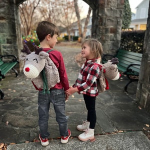 May include: Two children wearing reindeer-shaped backpacks, holding hands. The backpacks are gray with red noses and white accents. One child wears a red shirt and jeans, the other a plaid shirt and black pants. The scene is outdoors.
