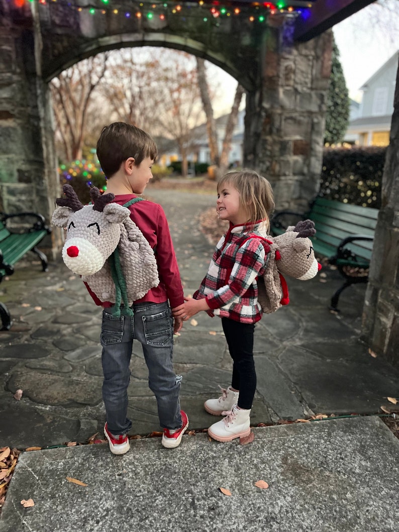 May include: Two children wearing reindeer-shaped backpacks, holding hands. The backpacks are gray with red noses and white accents. One child wears a red shirt and jeans, the other a plaid shirt and black pants. The scene is outdoors.