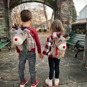 May include: Two children wearing reindeer-themed backpacks, holding hands, stand in front of a stone archway decorated with string lights. The backpacks are gray with brown antlers, red noses, and white accents. One child wears a red shirt and jeans, the other a plaid shirt and black leggings.