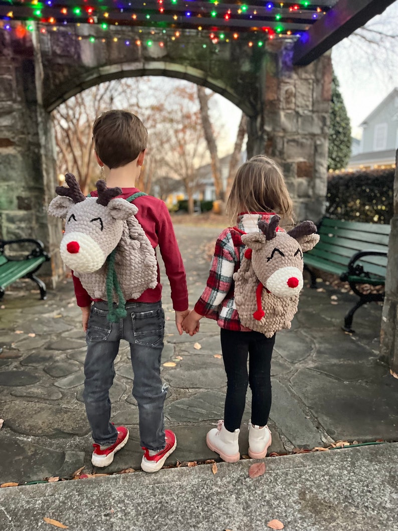 May include: Two children wearing reindeer-themed backpacks, holding hands, stand in front of a stone archway decorated with string lights. The backpacks are gray with brown antlers, red noses, and white accents. One child wears a red shirt and jeans, the other a plaid shirt and black leggings.
