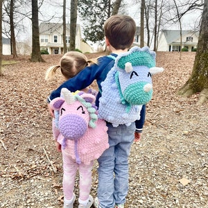 May include: Two children wearing crocheted animal backpacks. The girl is wearing a pink unicorn backpack and the boy is wearing a blue triceratops backpack. They are standing in a wooded area with trees and houses in the background.