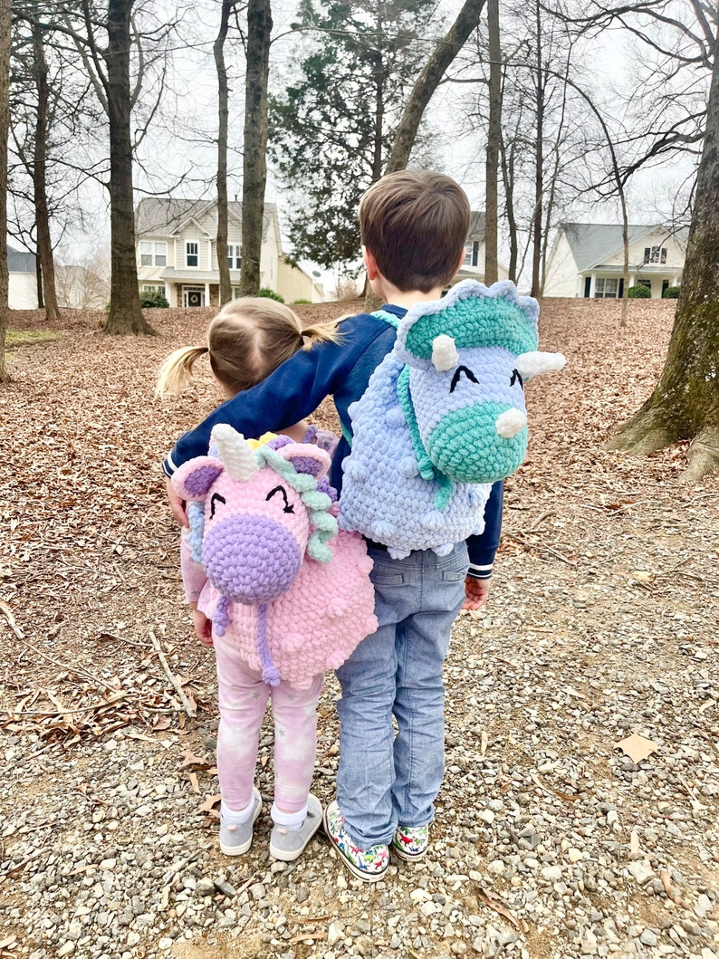 May include: Two children wearing crocheted animal backpacks. The girl is wearing a pink unicorn backpack and the boy is wearing a blue triceratops backpack. They are standing in a wooded area with trees and houses in the background.