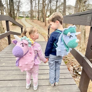 May include: Two children, a girl and a boy, are walking on a wooden bridge. The girl is wearing a pink and purple unicorn backpack and the boy is wearing a blue and green dinosaur backpack. They are both smiling and holding hands.
