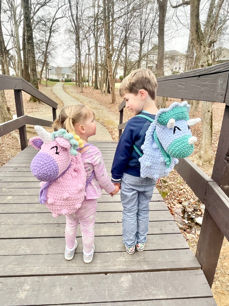 May include: Two children, a girl and a boy, are walking on a wooden bridge. The girl is wearing a pink and purple unicorn backpack and the boy is wearing a blue and green dinosaur backpack. They are both smiling and holding hands.