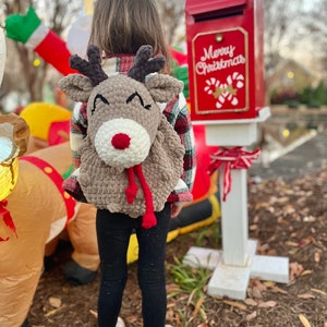 May include: A brown crocheted reindeer backpack with a white nose and red scarf is worn by a child. The backpack has antlers and a smiling face. The child is wearing black leggings and white boots. A red mailbox with "Merry Christmas" text is in the background.