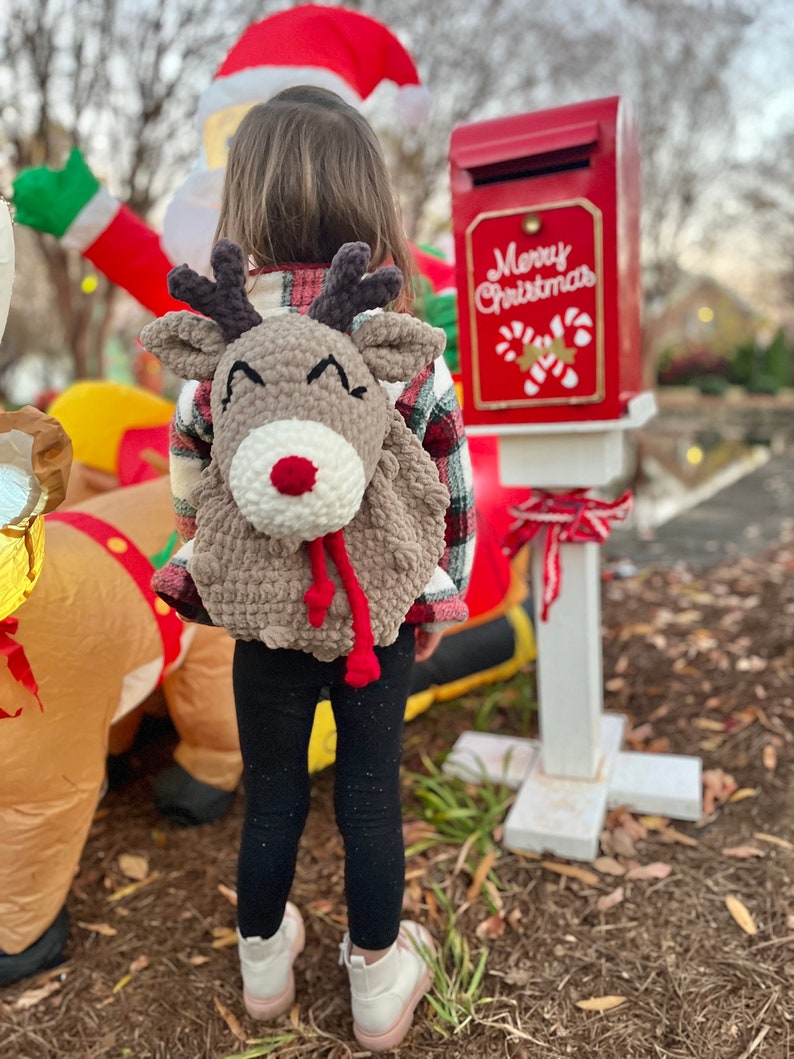 May include: A brown crocheted reindeer backpack with a white nose and red scarf is worn by a child. The backpack has antlers and a smiling face. The child is wearing black leggings and white boots. A red mailbox with "Merry Christmas" text is in the background.
