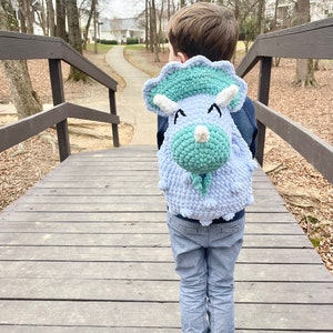 May include: A child wearing blue jeans is walking on a wooden bridge while wearing a light blue and green crocheted dinosaur backpack.