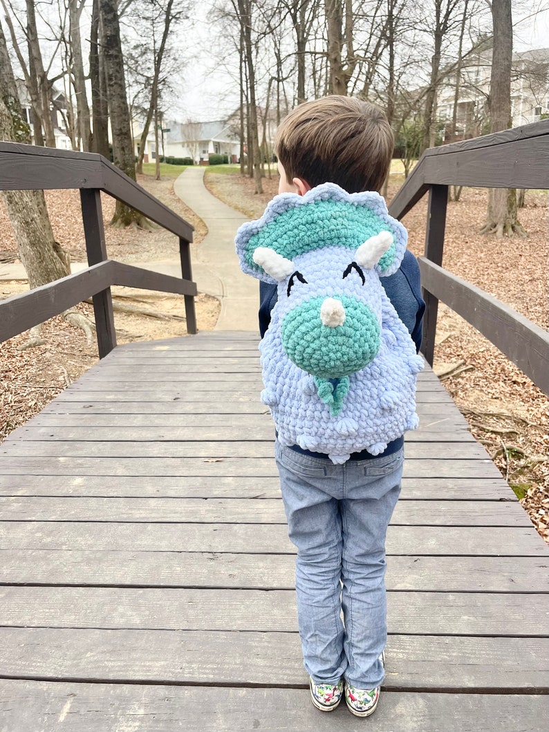 May include: A child wearing blue jeans is walking on a wooden bridge while wearing a light blue and green crocheted dinosaur backpack.