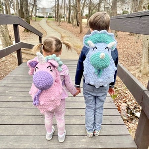 May include: Two children, one wearing a pink unicorn backpack and the other wearing a blue triceratops backpack, walk hand-in-hand on a wooden bridge.